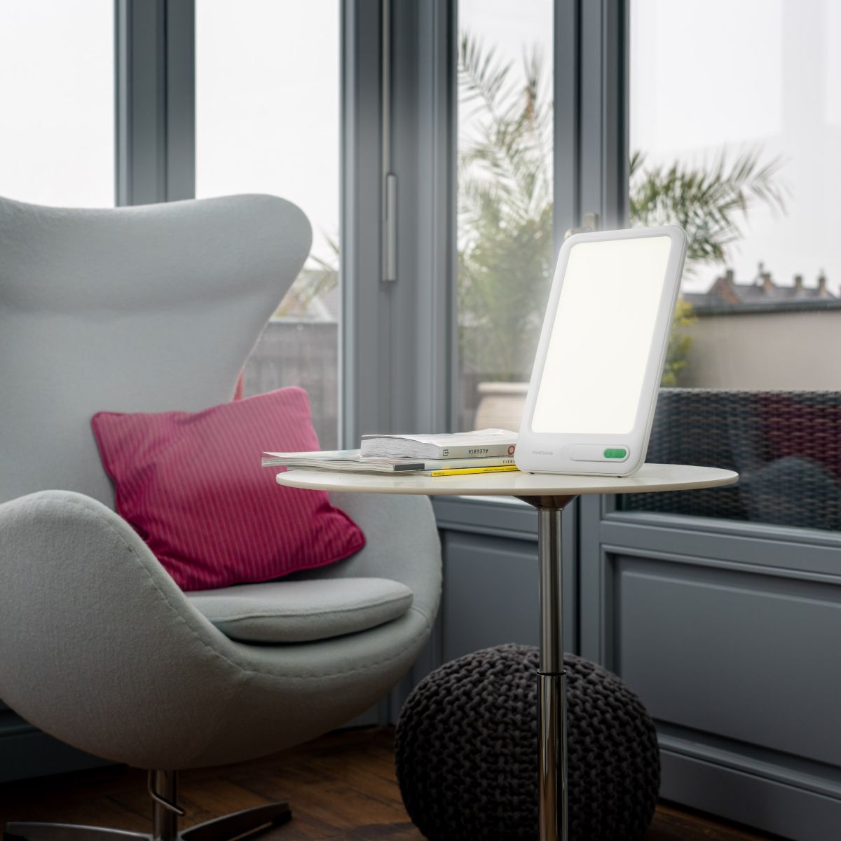 Eine eingeschaltete Tageslichtlampe steht auf einem kleinen Tisch neben einem Sessel und Büchern in einem hellen Wohnraum.